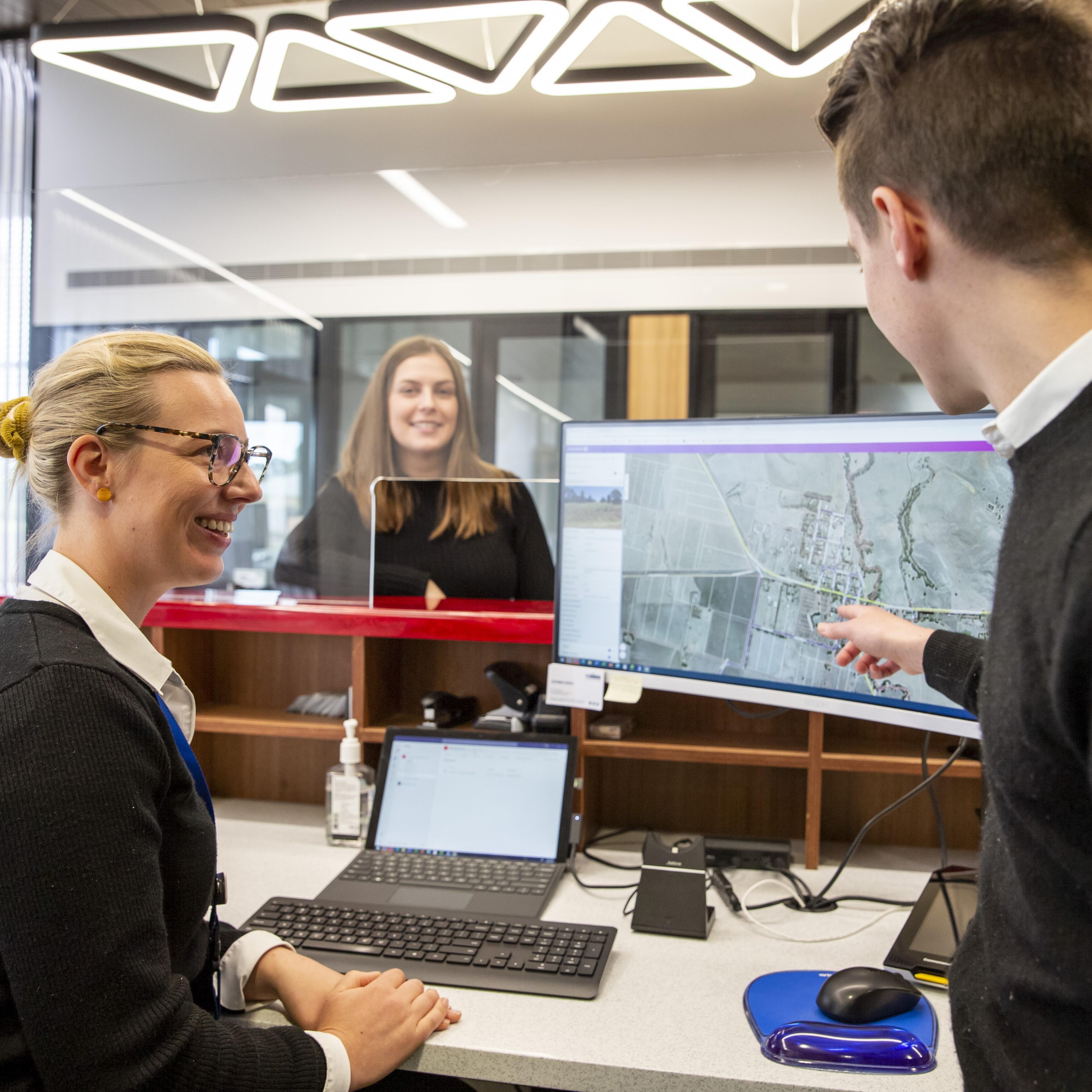 Three employees in an office one seated at a desk, another standing beside her. The third is watching from a distance. They are looking at a computer monitor and pointing at the content.